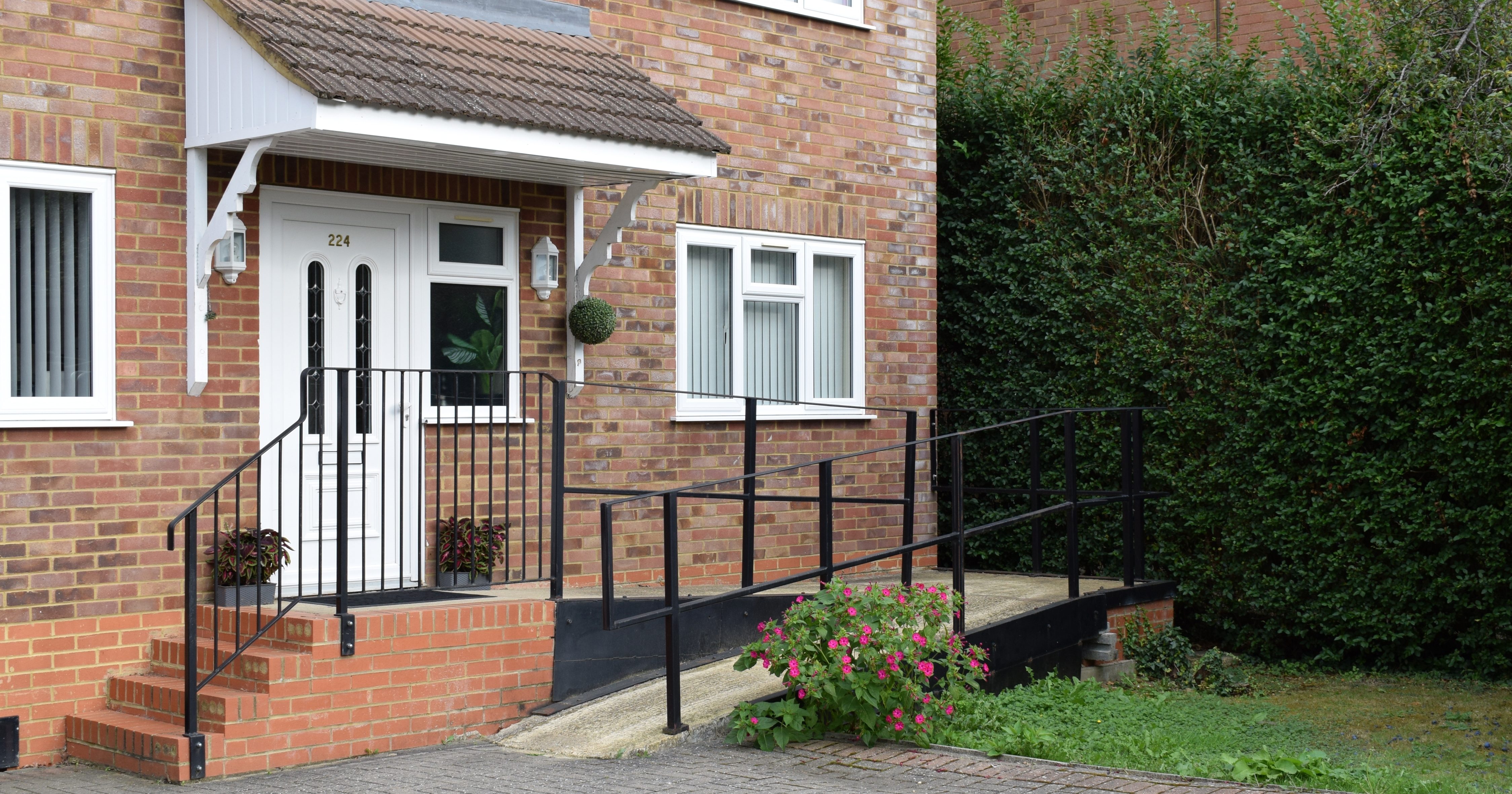 A brick house with white trim. There are steps and a ramp leading to the front door.  There is a big pot of pink flowers near the ramp.